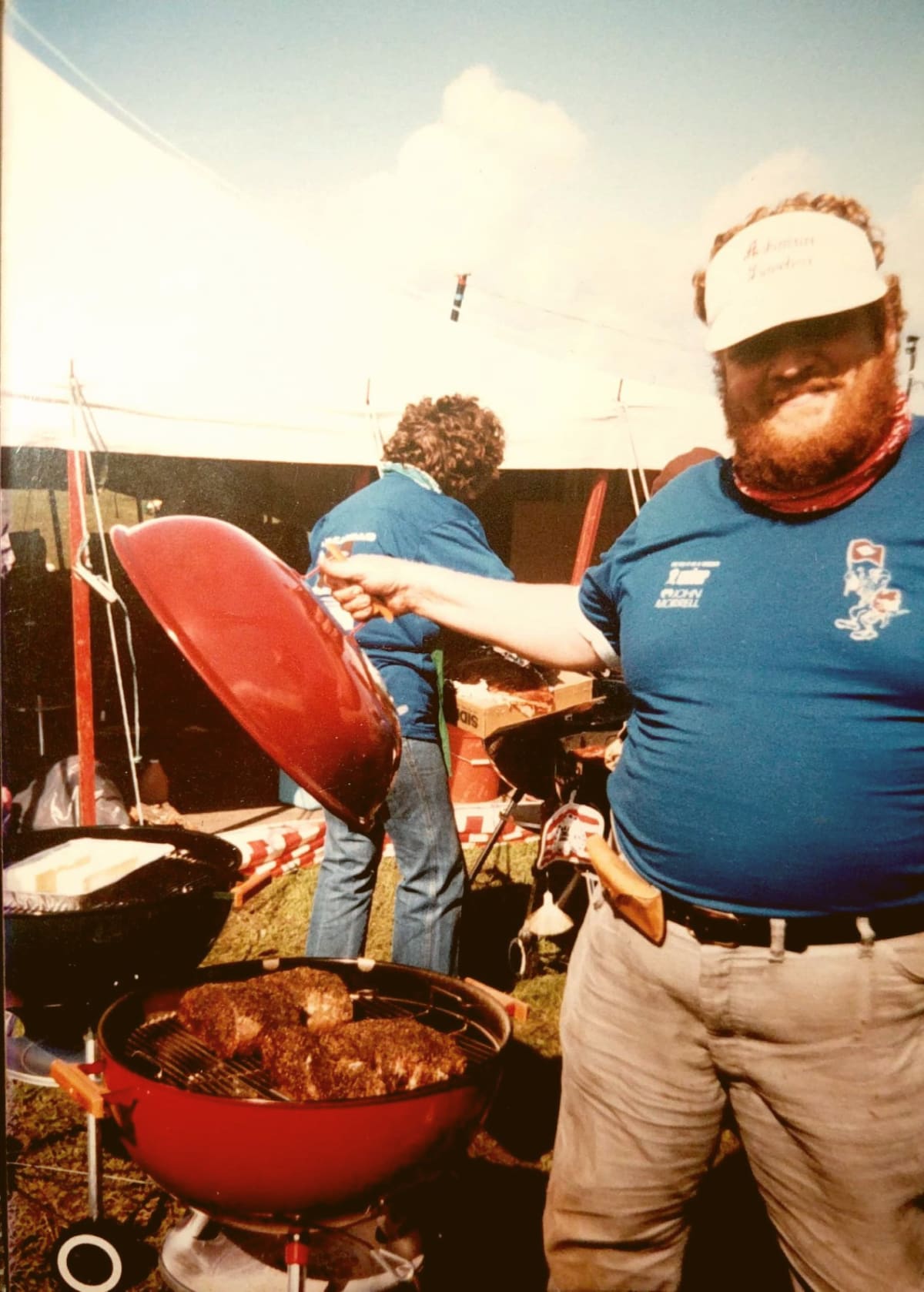 Jim Quessenberry, wearing his signature red hat, tenders a smoking chicken on a classic red grill.